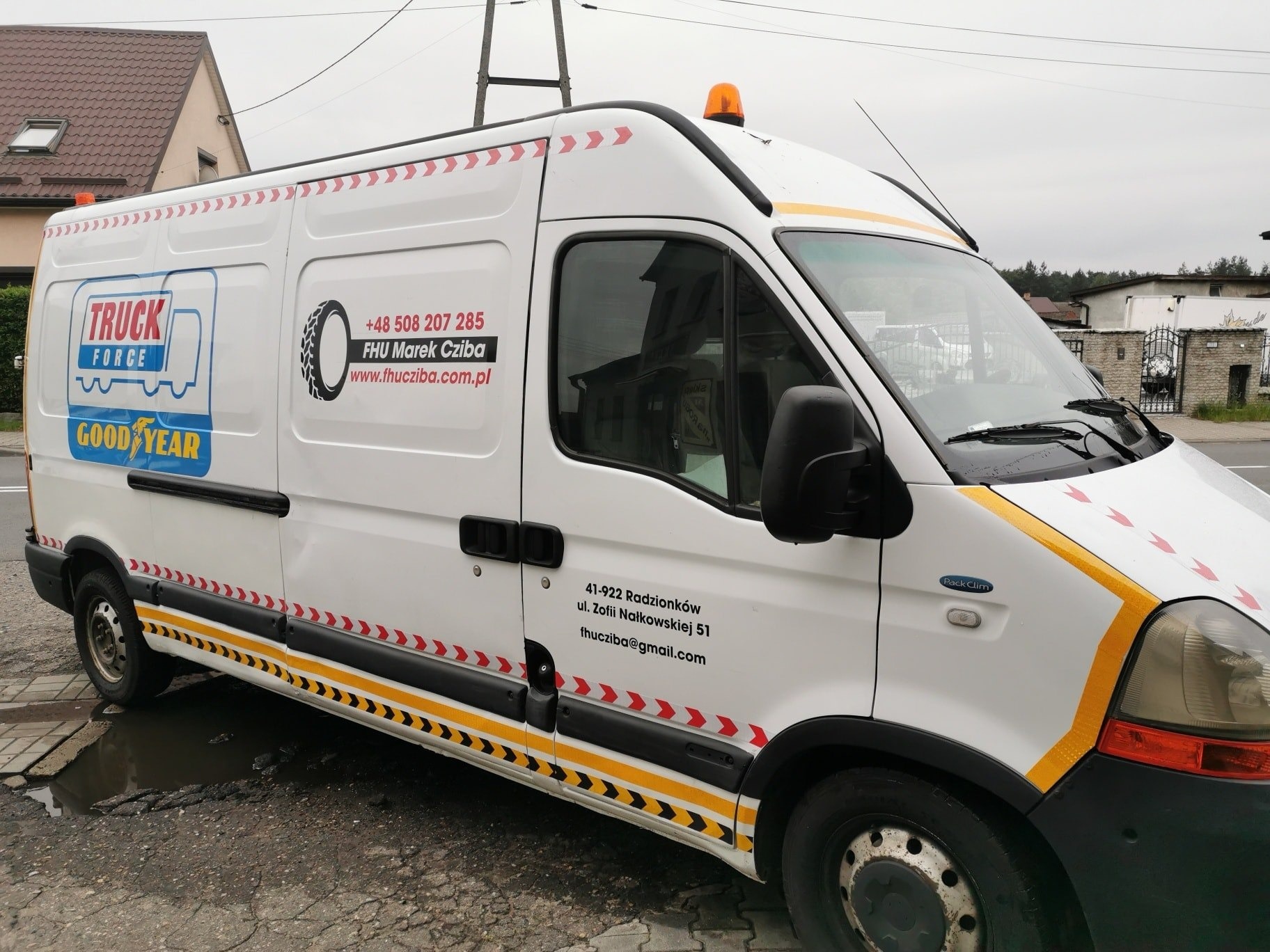 Biały samochód dostawczy z oklejonymi elementami graficznymi: logo firmy, dane kontaktowe i pasy ostrzegawcze w kolorach żółtym i czerwonym.
