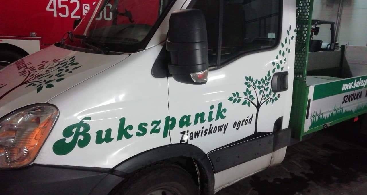 Biały samochód dostawczy z zielonymi naklejkami drzew i napisem 'Bukszpanik Żawiskowy ogród', zaparkowany w pomieszczeniu.