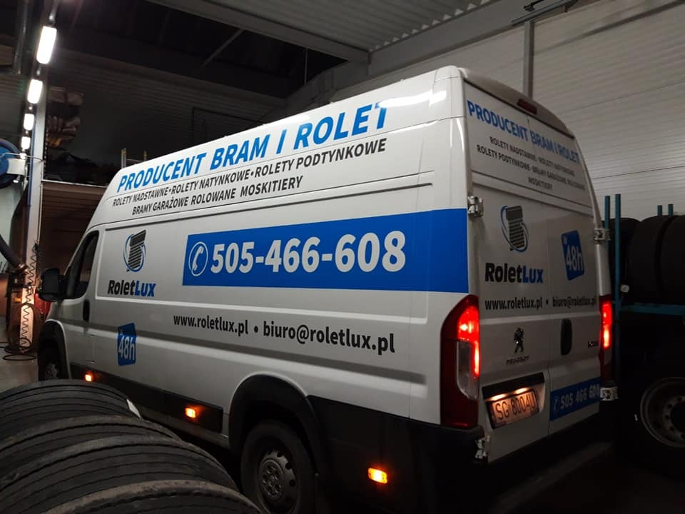 Biały samochód dostawczy z niebieskimi akcentami, oklejony reklamą producenta bram i rolet, zaparkowany w warsztacie, widoczne opony w tle.