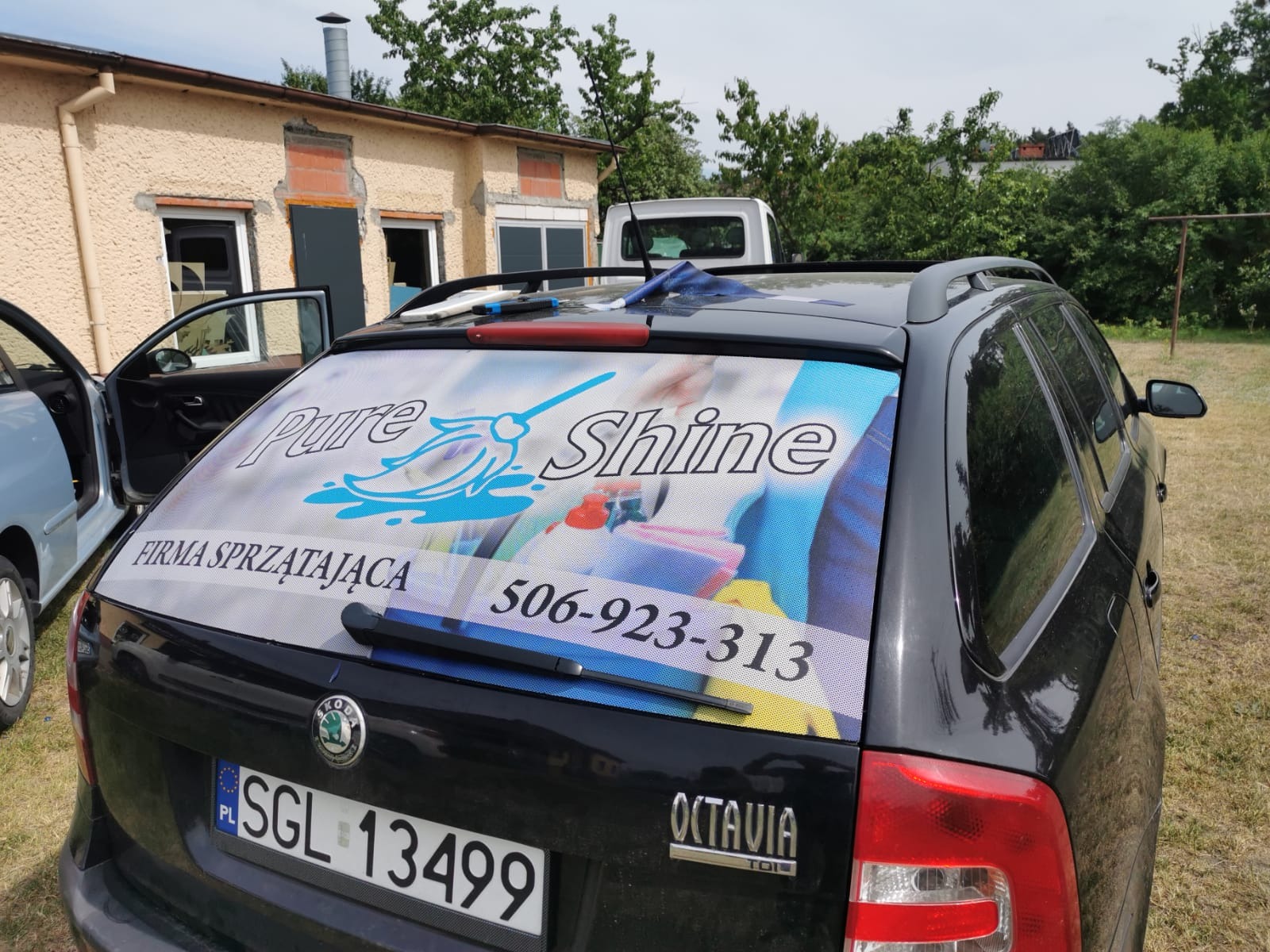 Czarny samochód Skoda Octavia z oklejoną tylną szybą reklamą firmy sprzątającej Pure Shine, zaparkowany na trawie przed budynkiem.