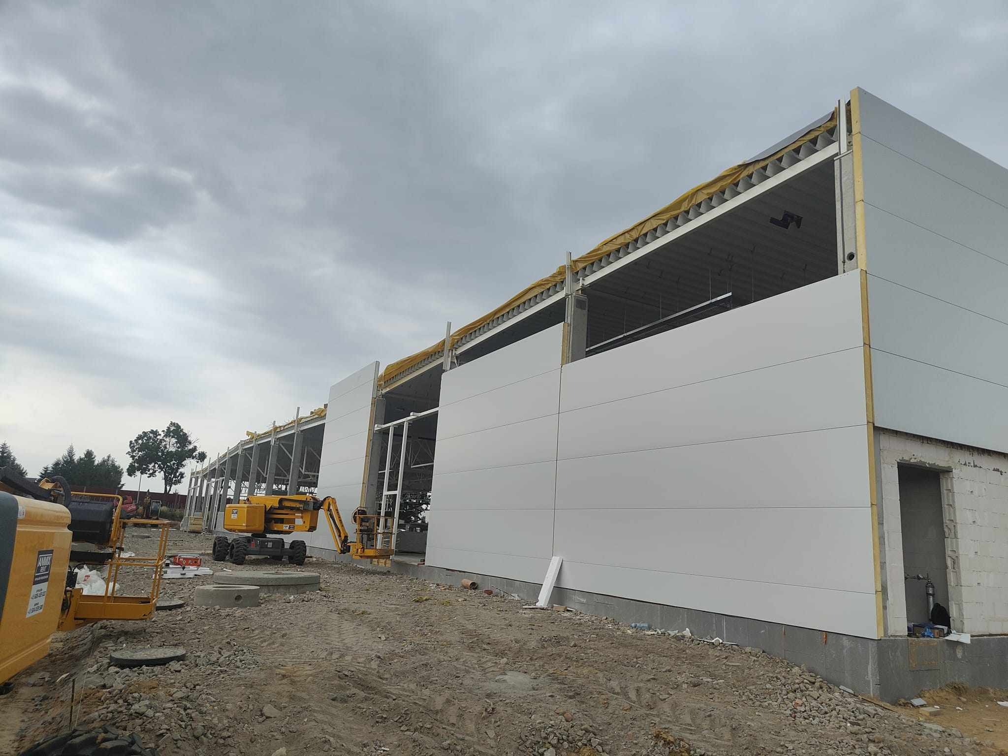 Wznoszenie hali z paneli warstwowych, widoczny szkielet konstrukcji i żółty podnośnik koszowy na placu budowy, pochmurne niebo.