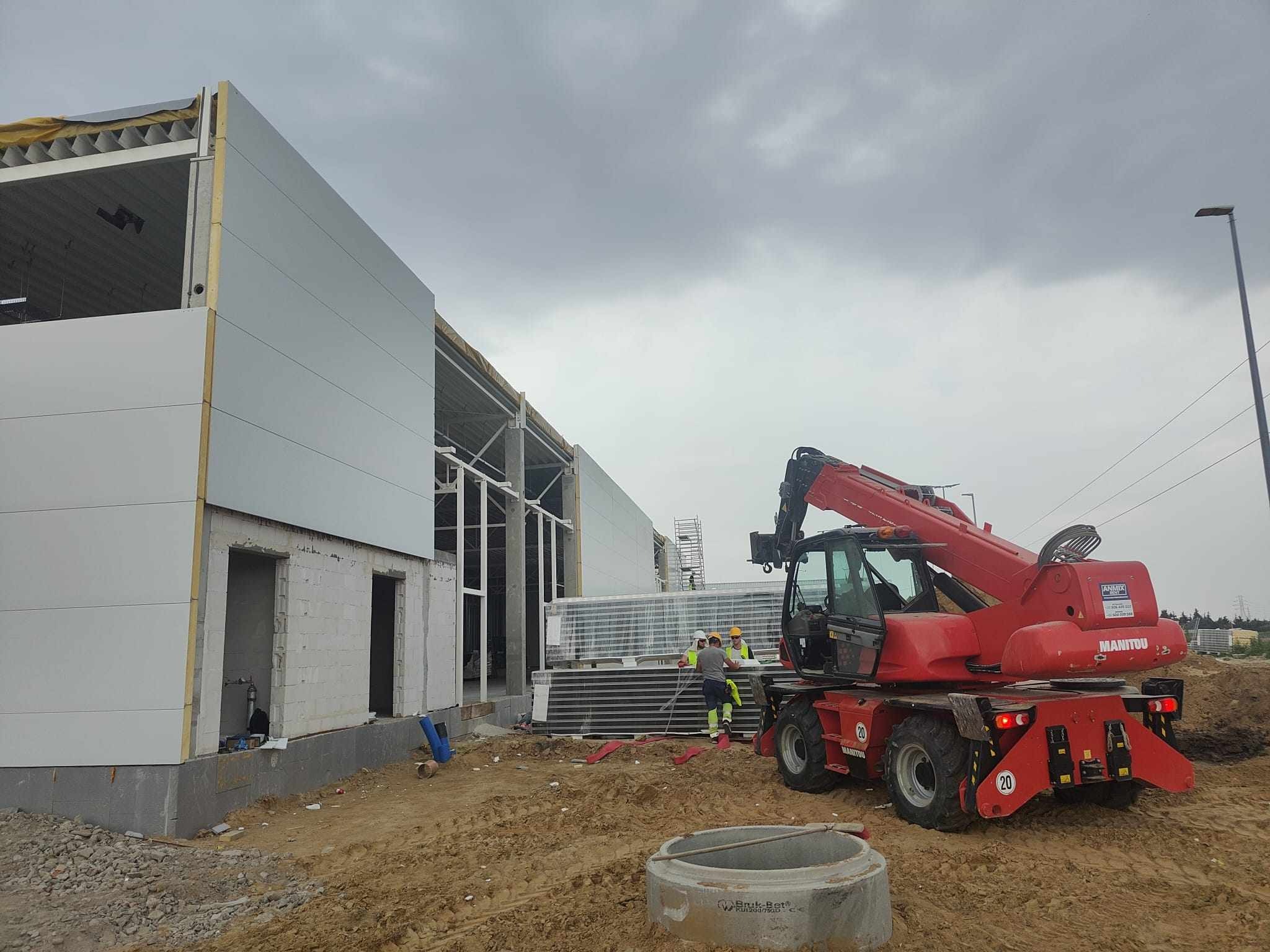 Montaż paneli ściennych na konstrukcji stalowej hali przemysłowej z użyciem czerwonego podnośnika teleskopowego Manitou; pracownicy w kaskach i kamizelkach odblaskowych przy panelach.