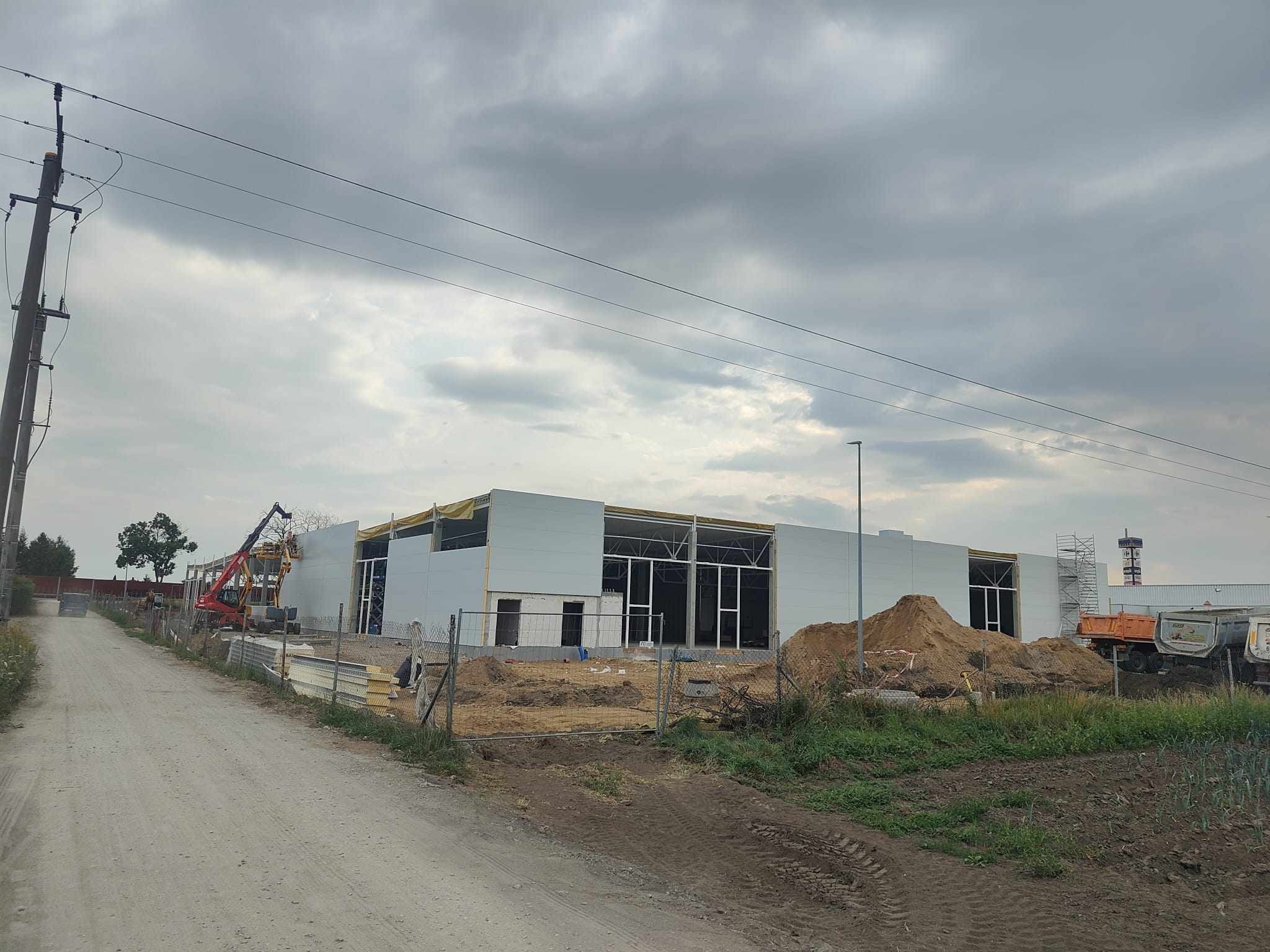 Wznoszenie hali: widok z zewnątrz na konstrukcję w trakcie montażu, z widocznym dźwigiem i stertami ziemi oraz częściowo zamontowanymi panelami ściennymi.