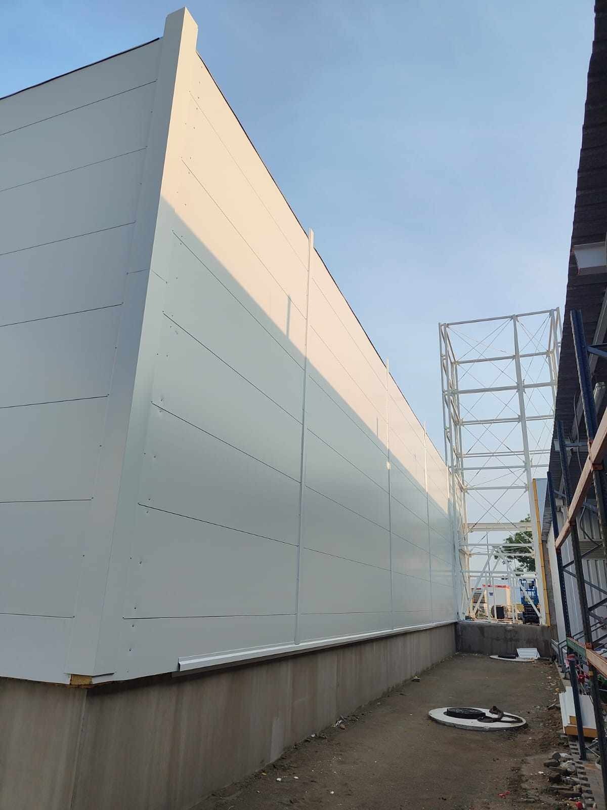 Fragment zewnętrznej ściany nowej hali z paneli warstwowych, widok z dołu na tle niebieskiego nieba i konstrukcji wsporczej.