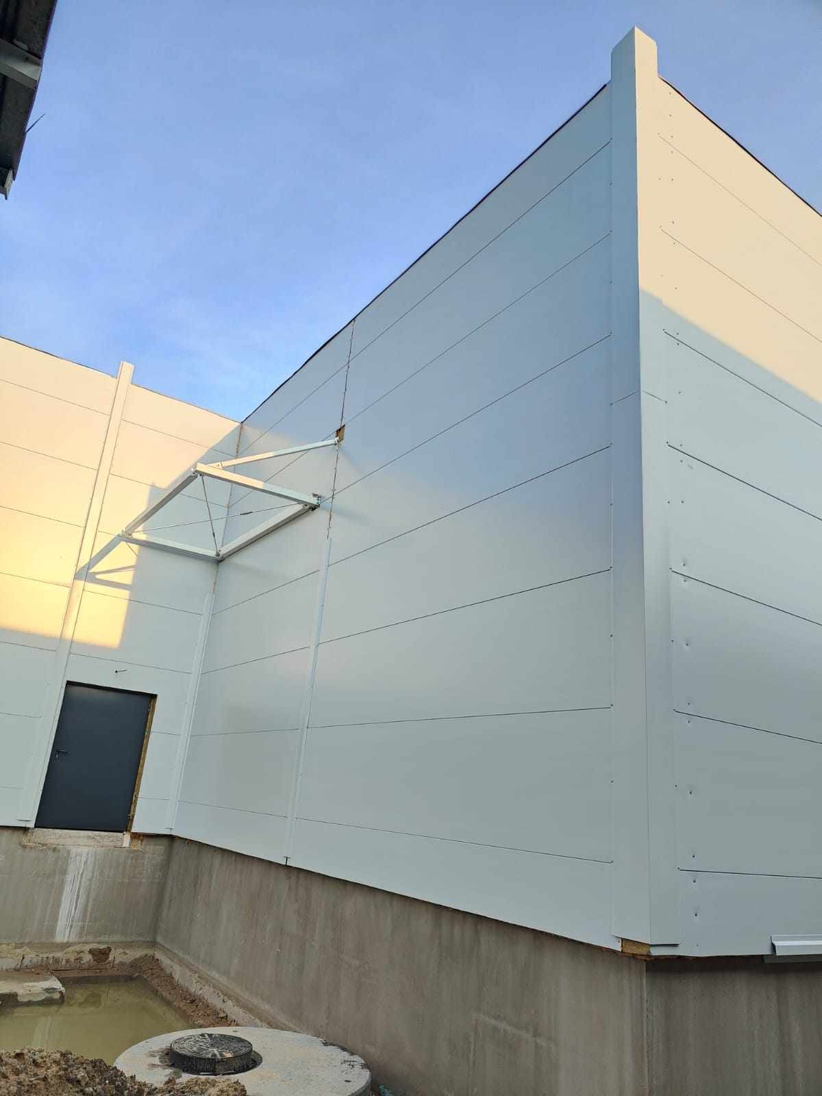 Zewnętrzny widok hali z paneli warstwowych w kolorze jasnoszarym, z widocznym wejściem i elementami konstrukcyjnymi na tle błękitnego nieba.