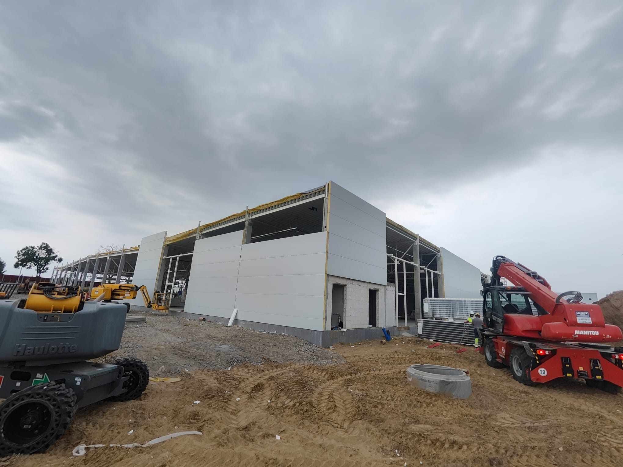 Budowa hali z widocznymi elementami konstrukcji stalowej, maszyny budowlane Haulotte i Manitou na placu budowy, pochmurne niebo.