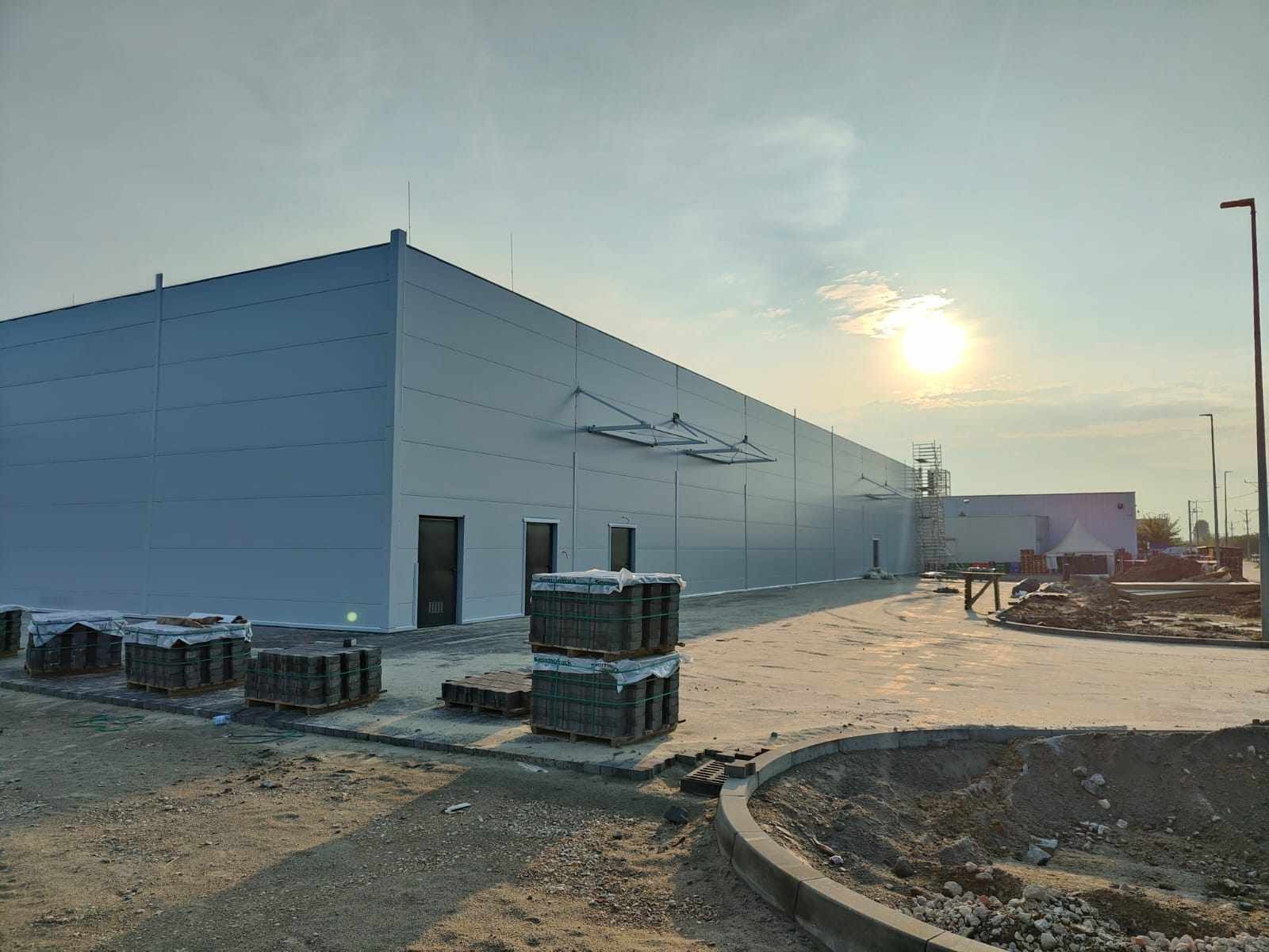 Nowoczesna hala magazynowa w trakcie budowy, widoczne elementy konstrukcyjne i materiały budowlane, plac budowy w promieniach zachodzącego słońca.