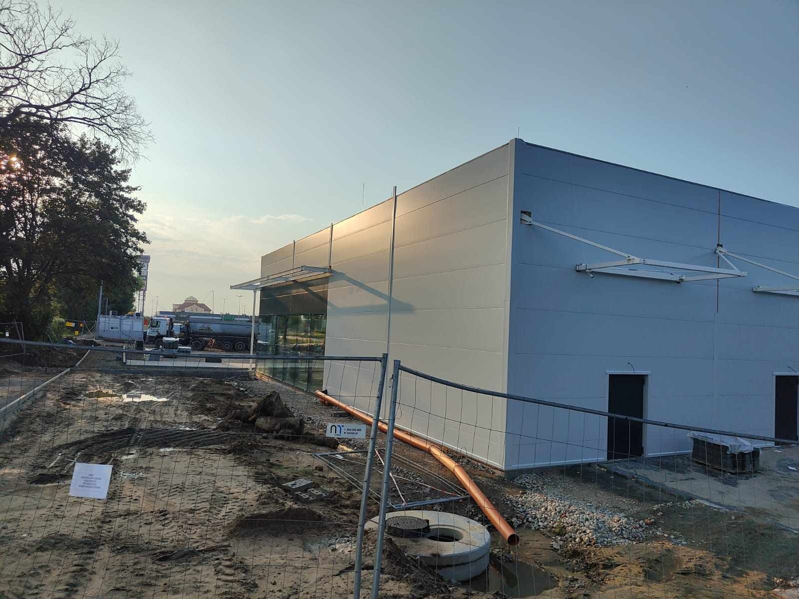 Nowoczesna hala przemysłowa z szarej blachy trapezowej w trakcie budowy, widoczne elementy konstrukcyjne, teren budowy zabezpieczony siatką, w tle zaparkowana cysterna.