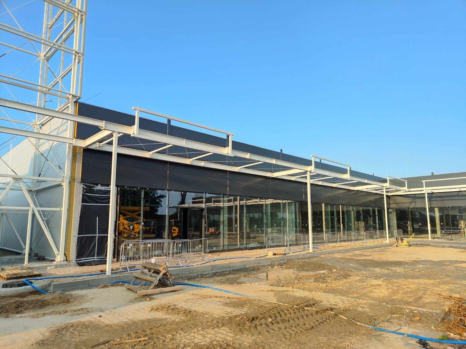 Wznoszona hala z elementami konstrukcji stalowej i dużymi przeszkleniami, widoczny plac budowy z rusztowaniami i materiałami budowlanymi.
