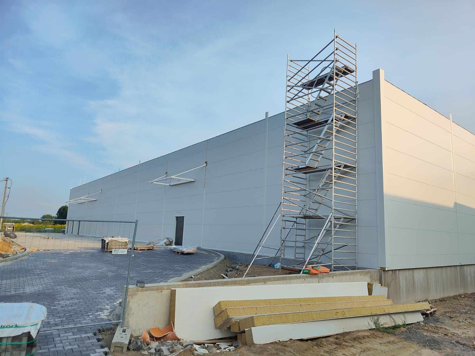 Szara hala przemysłowa w trakcie budowy, widoczne rusztowanie, materiały izolacyjne i kostka brukowa na placu budowy pod błękitnym niebem.