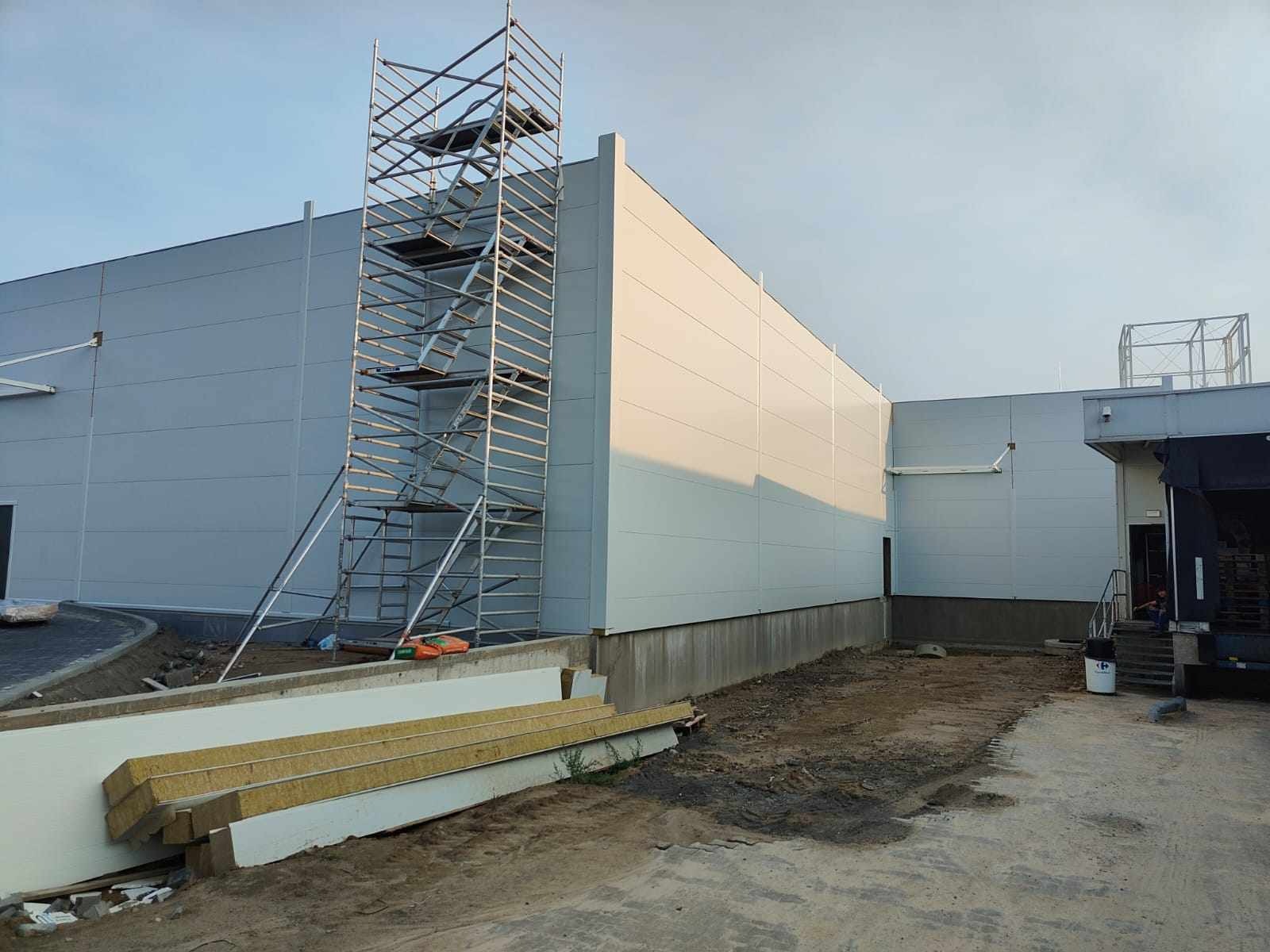 Montaż paneli ściennych na hali przemysłowej, widoczne rusztowanie i przygotowane płyty warstwowe.