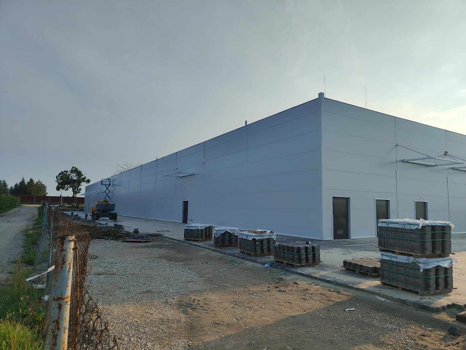 Szara hala magazynowa w trakcie budowy z paletami kostki brukowej ułożonymi wzdłuż ściany i podnośnikiem koszowym w tle.