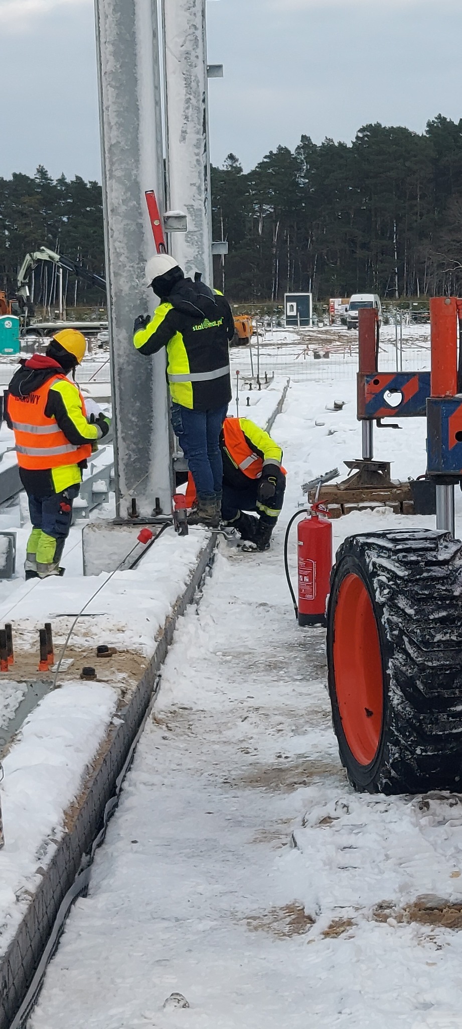 Montaż stalowych słupów konstrukcyjnych na śnieżnym placu budowy, pracownicy w odblaskowych kamizelkach sprawdzają pion za pomocą poziomicy, w tle las i sprzęt budowlany.