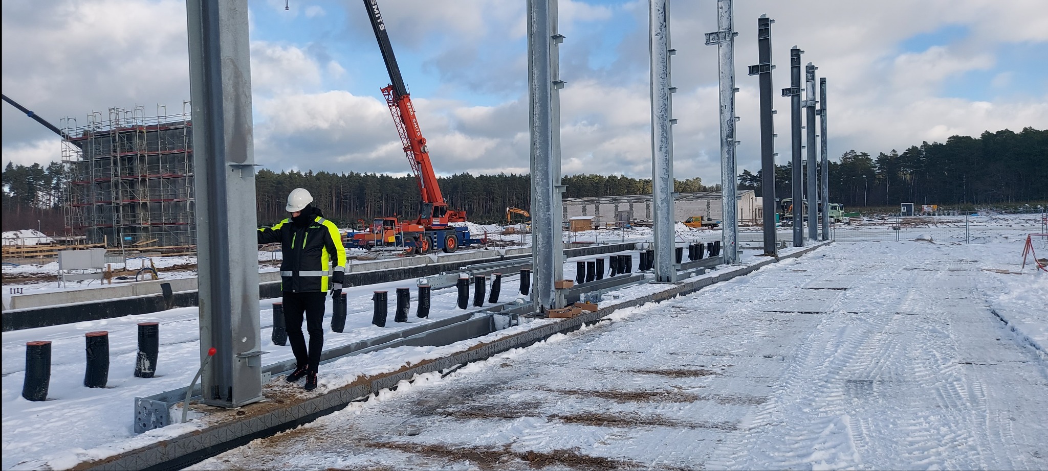 Montaż stalowej konstrukcji hali w zimowej scenerii, widoczny pracownik w kasku i odblaskowej kurtce, żuraw budowlany i inne maszyny w tle.
