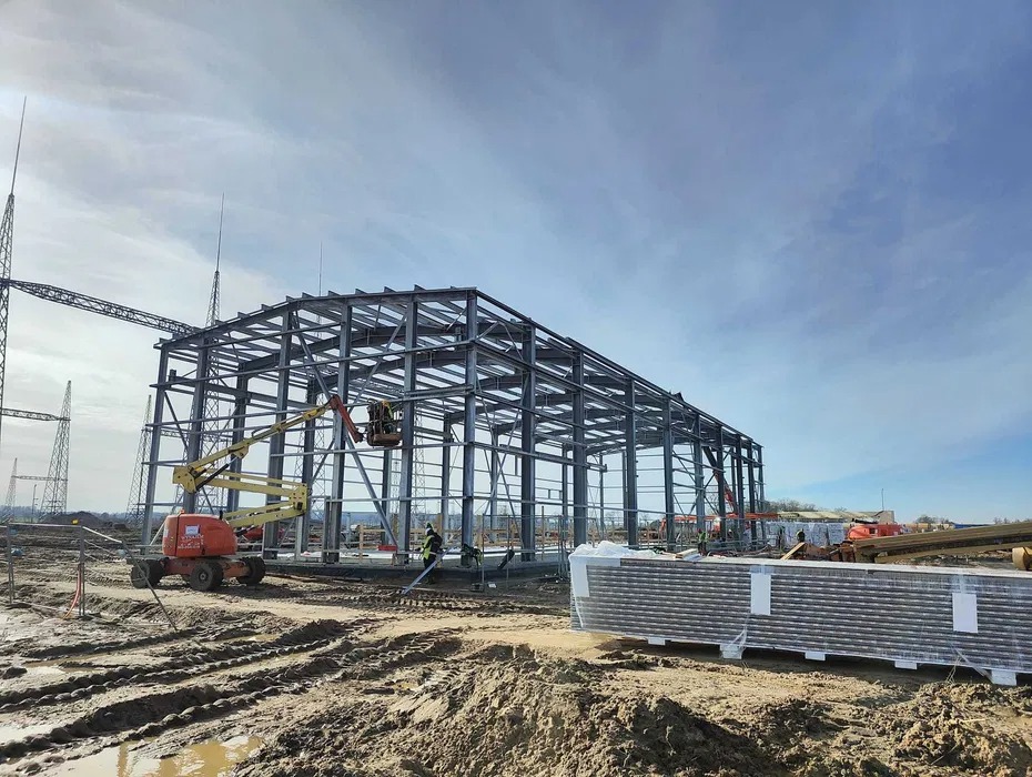 Stalowa konstrukcja hali w trakcie montażu, widoczny podnośnik koszowy i materiały izolacyjne na ziemi.