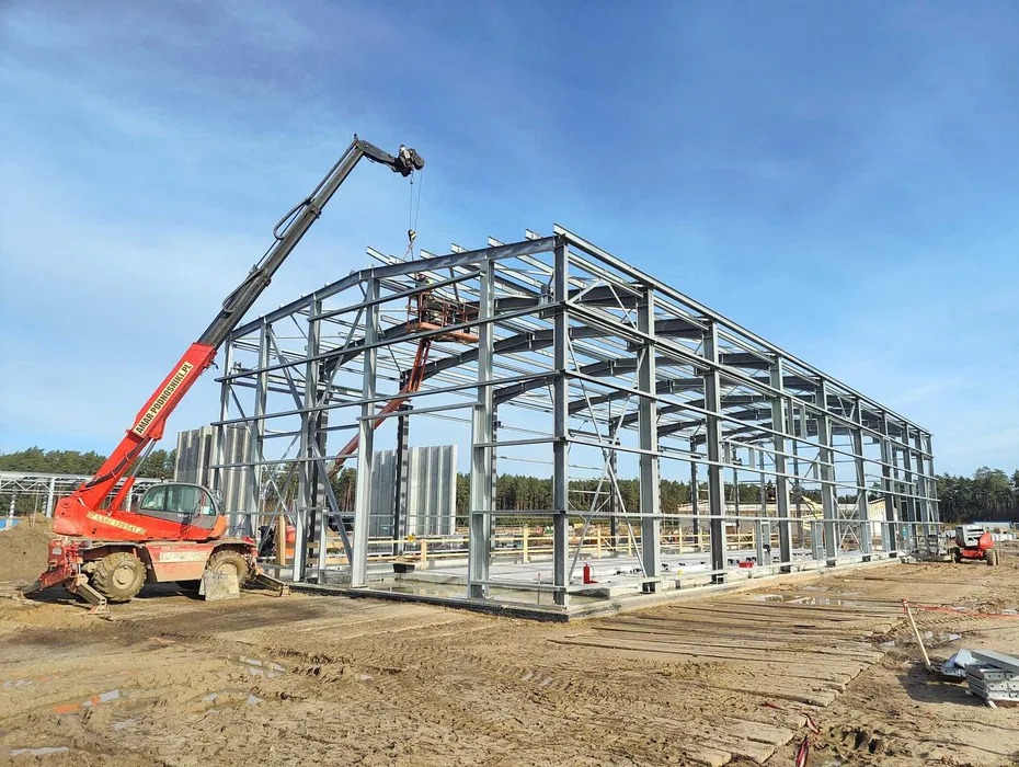 Montaż konstrukcji stalowej hali z użyciem podnośnika teleskopowego na tle błękitnego nieba i lasu.