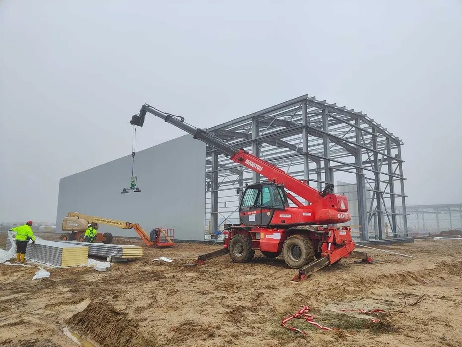 Montaż paneli ściennych hali stalowej przy użyciu przyssawki podwieszonej do czerwonego teleskopowego podnośnika Manitou; w tle szkielet konstrukcji i podnośnik koszowy.