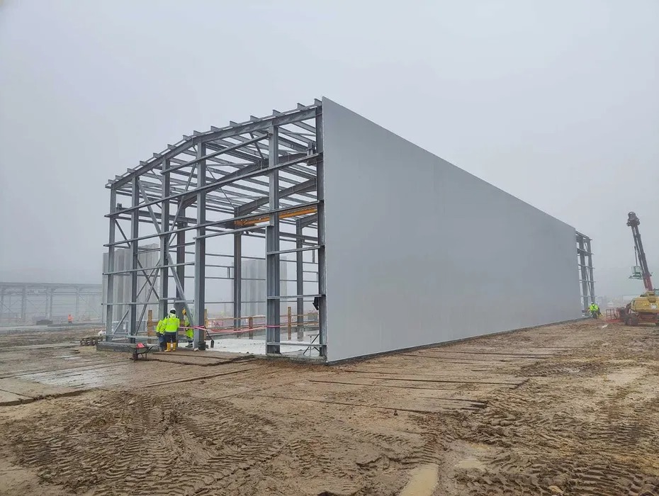 Budowa hali stalowej w Warszawie w trakcie montażu paneli ściennych; widoczna konstrukcja szkieletowa, częściowo obudowana, oraz pracownicy w odblaskowych kamizelkach na błotnistym terenie.