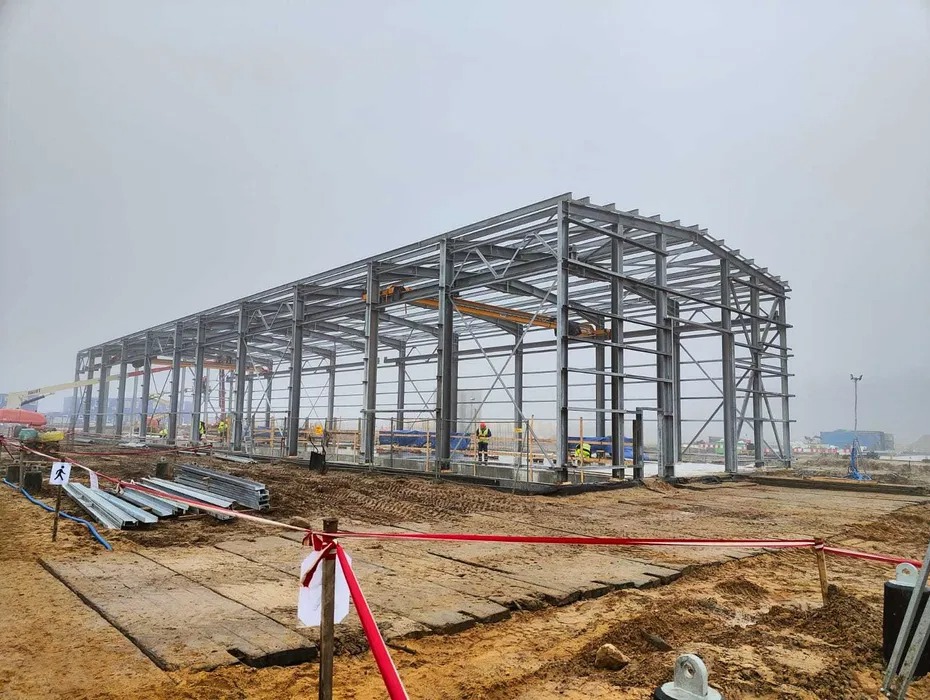 Stalowa konstrukcja hali w trakcie montażu, widoczny dźwig pomostowy i pracownicy w kamizelkach odblaskowych na tle pochmurnego nieba.