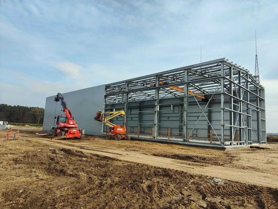 Montaż stalowej konstrukcji hali z użyciem podnośników teleskopowych na placu budowy, widoczne elementy obudowy i szkielet konstrukcyjny.