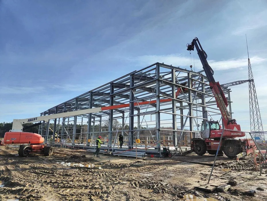 Montaż szkieletu stalowego hali z użyciem dźwigu Manitou i podnośnika JLG Lift, widoczni pracownicy w kamizelkach odblaskowych, teren budowy w trakcie prac.