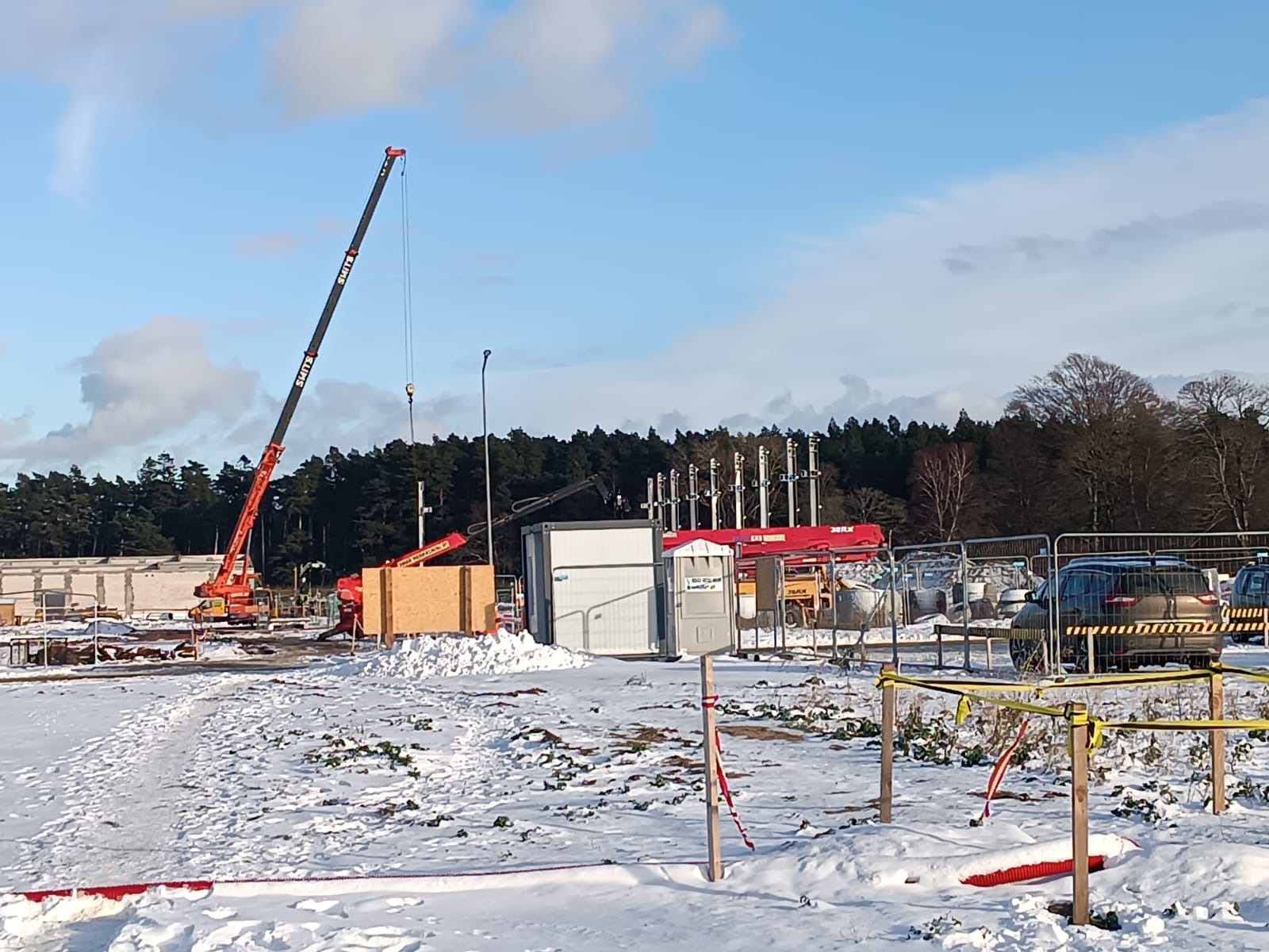Plac budowy w zimowej scenerii: czerwony dźwig, elementy konstrukcyjne, przenośna toaleta i zasypane śniegiem podłoże.