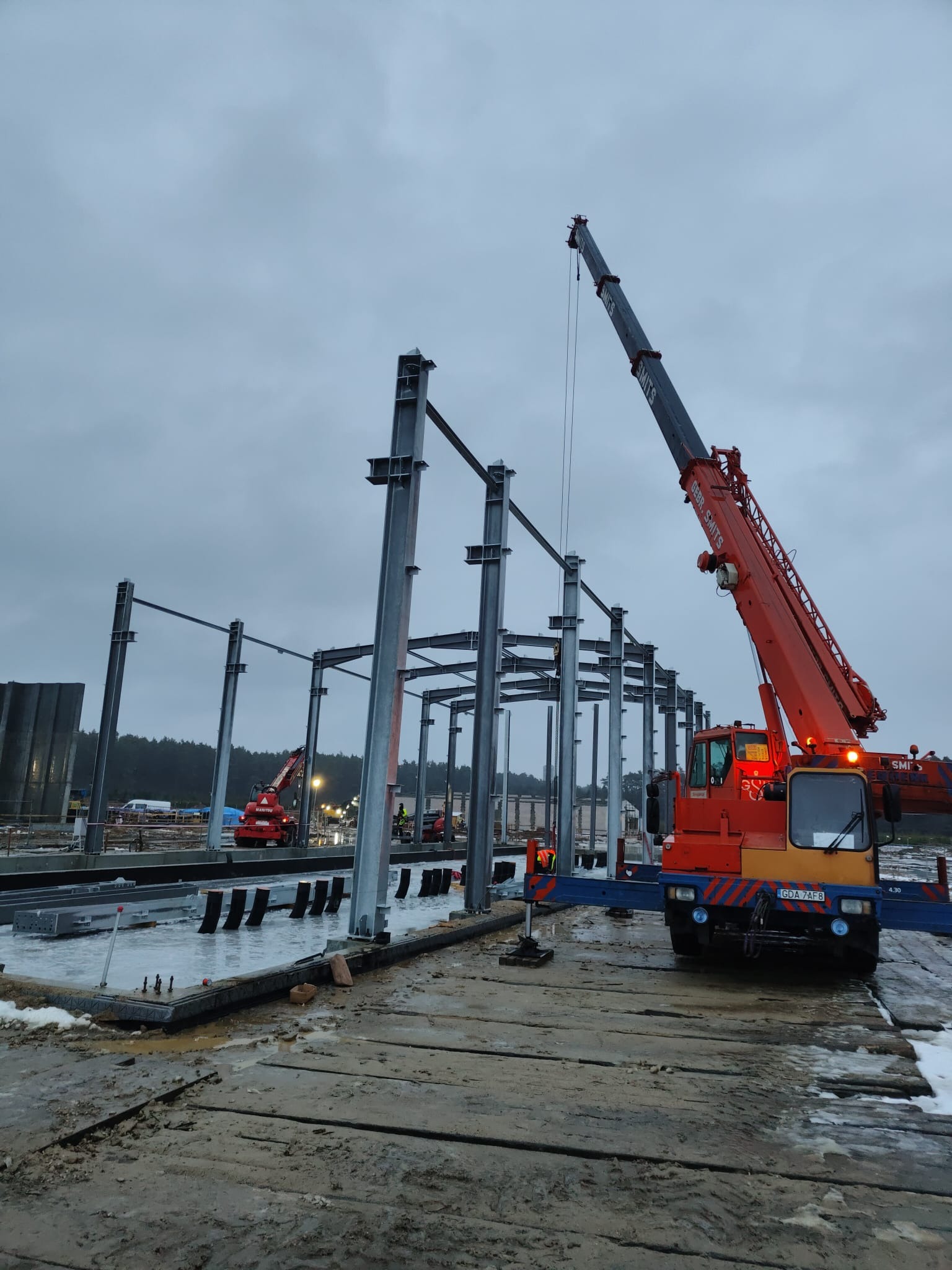 Montaż stalowej konstrukcji hali z użyciem żurawia samojezdnego Gebr. Smits pod pochmurnym niebem.