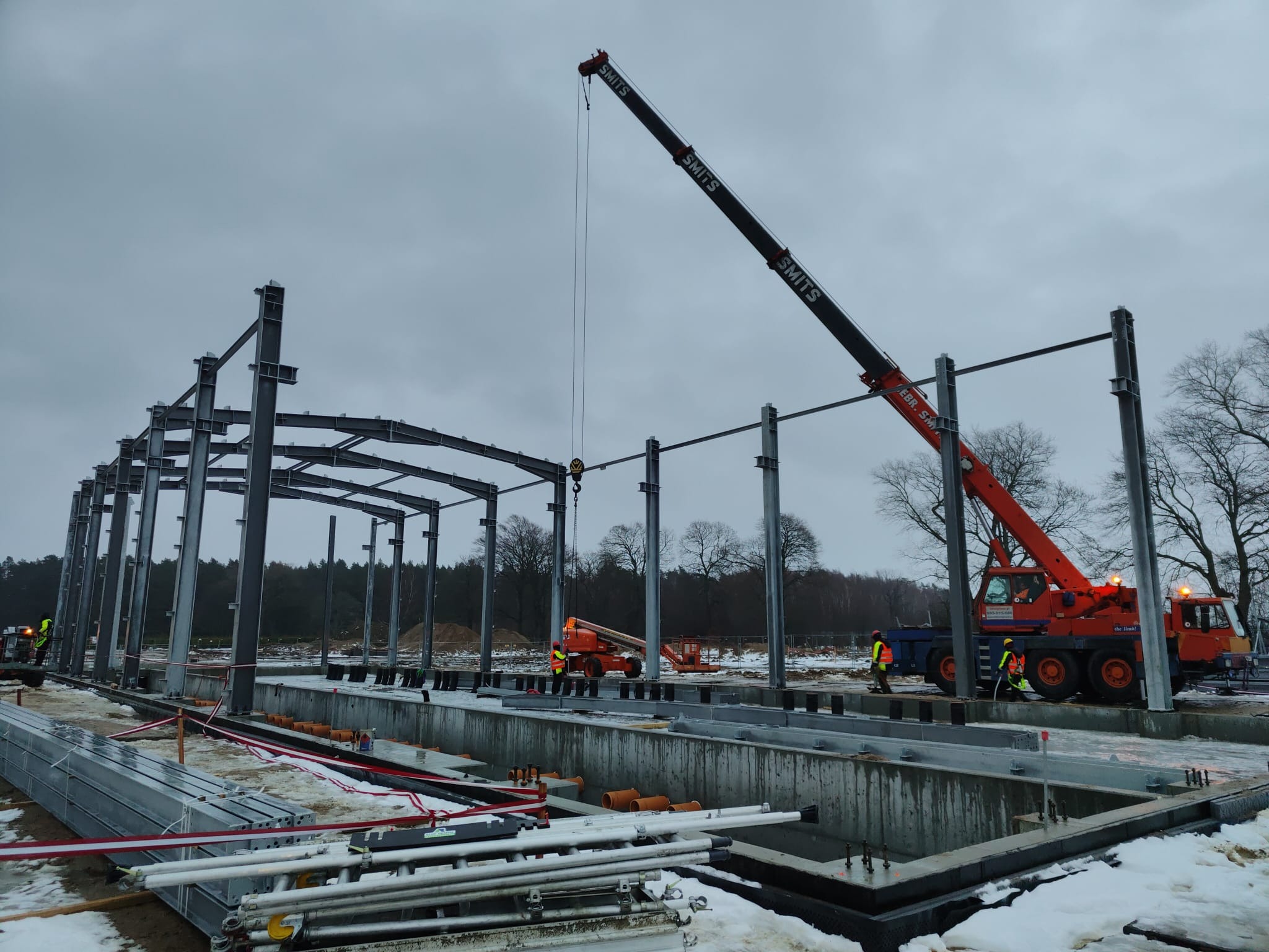 Montaż stalowej konstrukcji hali z użyciem dźwigu SMITS na tle pochmurnego nieba i zimowego krajobrazu.