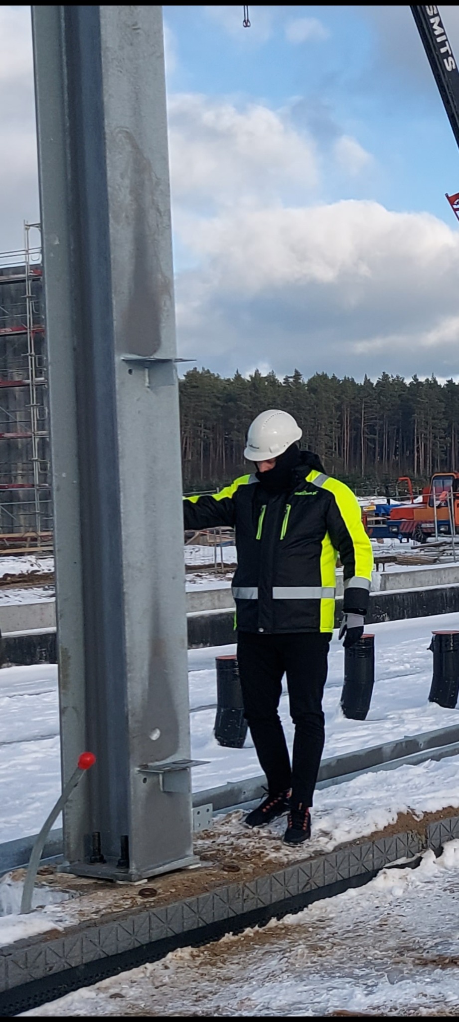 Pracownik w odblaskowej kurtce i kasku przy metalowej kolumnie na placu budowy hali, w tle widoczne rusztowania i dźwig.