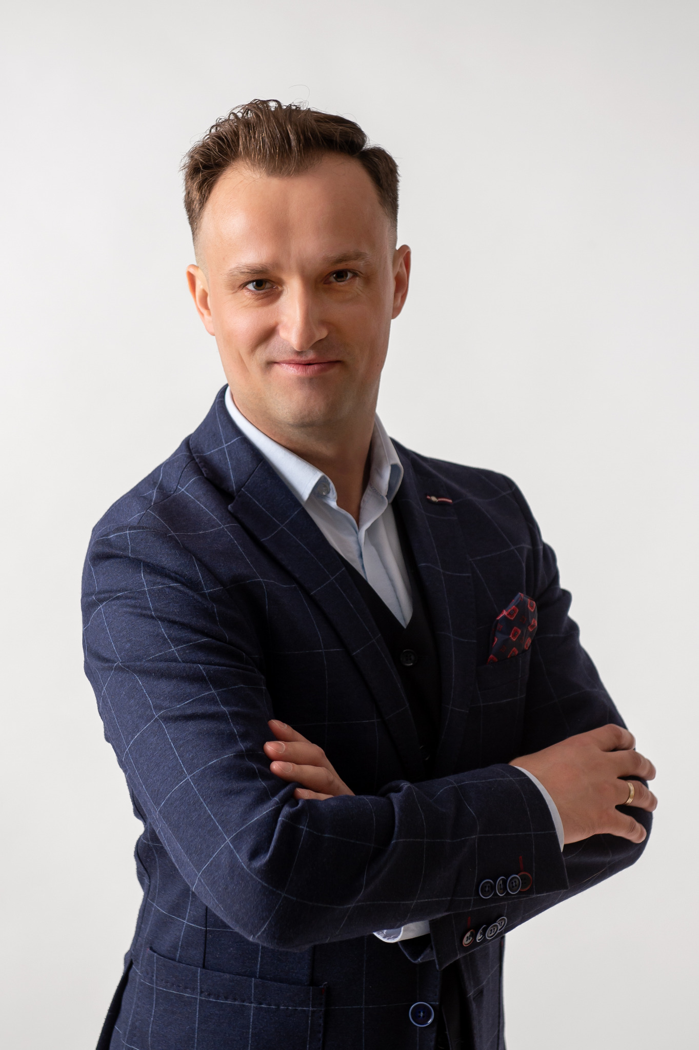 Portret uśmiechniętego mężczyzny w granatowym garniturze w kratę, z chusteczką w brustaszy, pozującego na jasnym tle z założonymi rękami.