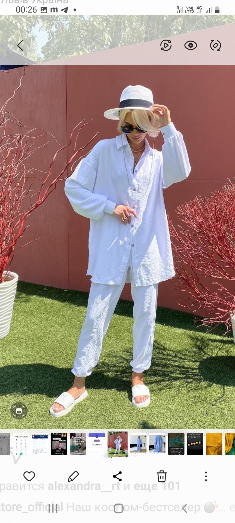 Kobieta w białym, luźnym komplecie odzieżowym (koszula i spodnie) oraz kapeluszu, stojąca na sztucznej trawie przed ścianą w kolorze terakoty z dekoracyjnymi czerwonymi gałęziami w donicach.