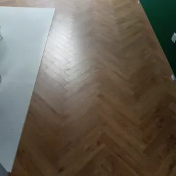 Ułożona jodełka z paneli podłogowych w odcieniach brązu, widoczny fragment białej ściany i ciemnozielonej ściany w tle.