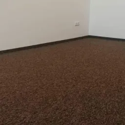 Brązowa wykładzina dywanowa z ciemnymi listwami przypodłogowymi w rogu pokoju o białych ścianach, widoczne gniazdko elektryczne.