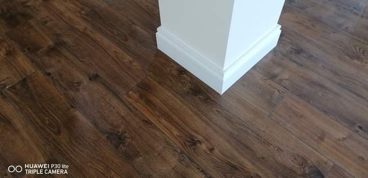 Ciemnobrązowe panele podłogowe ułożone przy białej listwie przypodłogowej, imitujące drewno z widocznym usłojeniem.