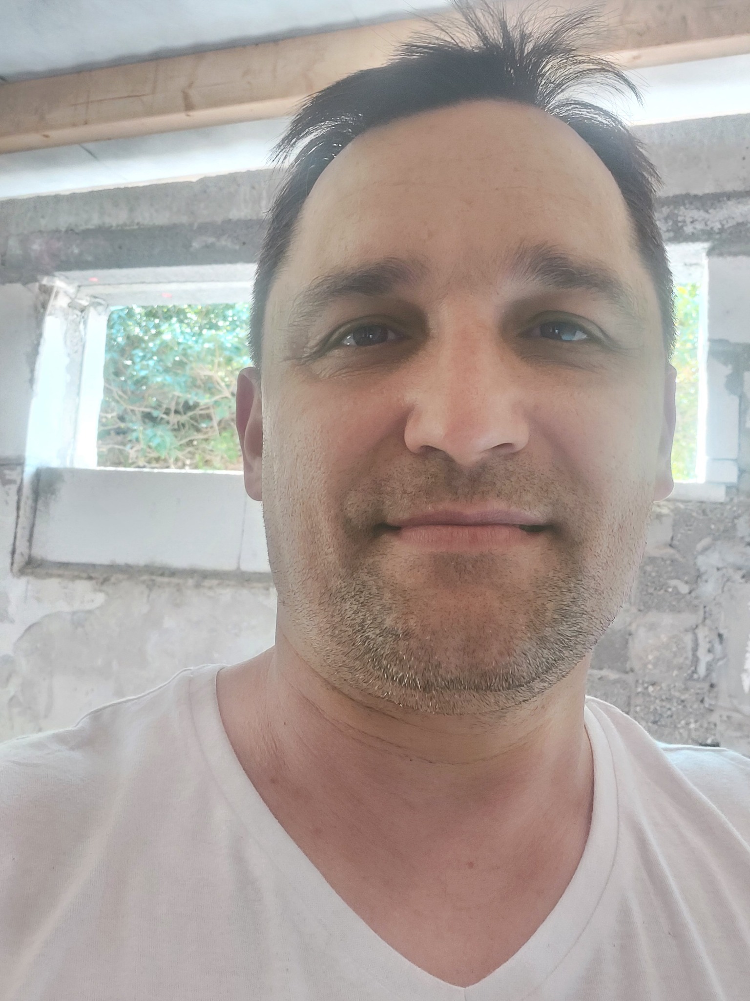 Portret mężczyzny w białej koszulce na tle surowego wnętrza z oknem, widoczne ślady prac budowlanych na ścianach.
