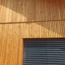 Drewniana elewacja budynku z jasnego drewna, widoczne pionowe deski i fragment okna z żaluzjami.