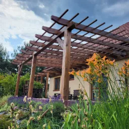 Drewniana konstrukcja z belek nad tarasem, widok od strony ogrodu z kwitnącymi liliowcami i lawendą na pierwszym planie, pochmurne niebo w tle.