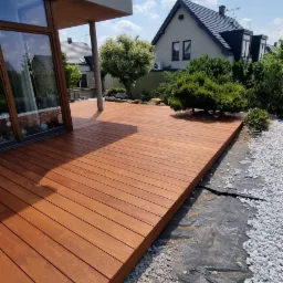 Nowoczesny taras z desek o ciepłej, brązowej barwie, przylegający do budynku z dużym oknem, otoczony kamieniami i zielenią w słoneczny dzień.