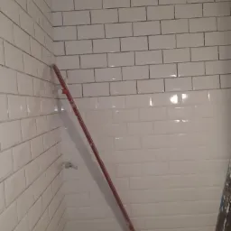 Świeżo ułożone białe, prostokątne płytki z ciemnoszarą fugą na ścianach łazienki, z widocznymi śladami prac wykończeniowych i częścią drabiny malarskiej.