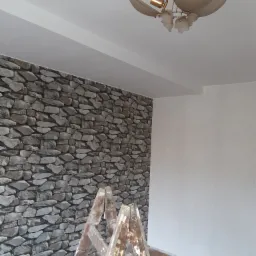 Ściana wyłożona kamieniem dekoracyjnym w Mysłowicach, widoczna drabina malarska i sufit z lampą w kształcie kwiatu.