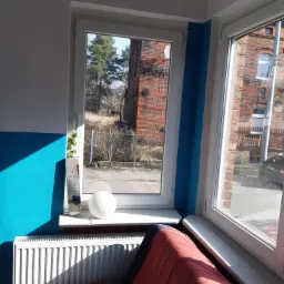 Dwa białe okna PCV z widokiem na ceglany budynek i drzewa, ściana pomalowana na biało i niebiesko, biały grzejnik pod oknem, na parapecie dekoracja z rośliną i białą kulą.