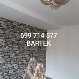 Ściana wykończona kamieniem dekoracyjnym w szarościach, widoczna drabina malarska, numer telefonu i imię wykonawcy naniesione cyfrowo na zdjęcie.