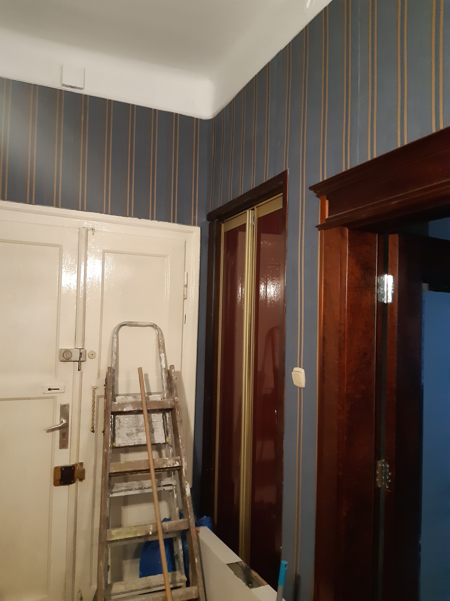 Pokój w trakcie odświeżania: drabina oparta o białe drzwi na tle ściany w szaro-złote pasy, obok ciemne, drewniane drzwi przesuwne i futryna.