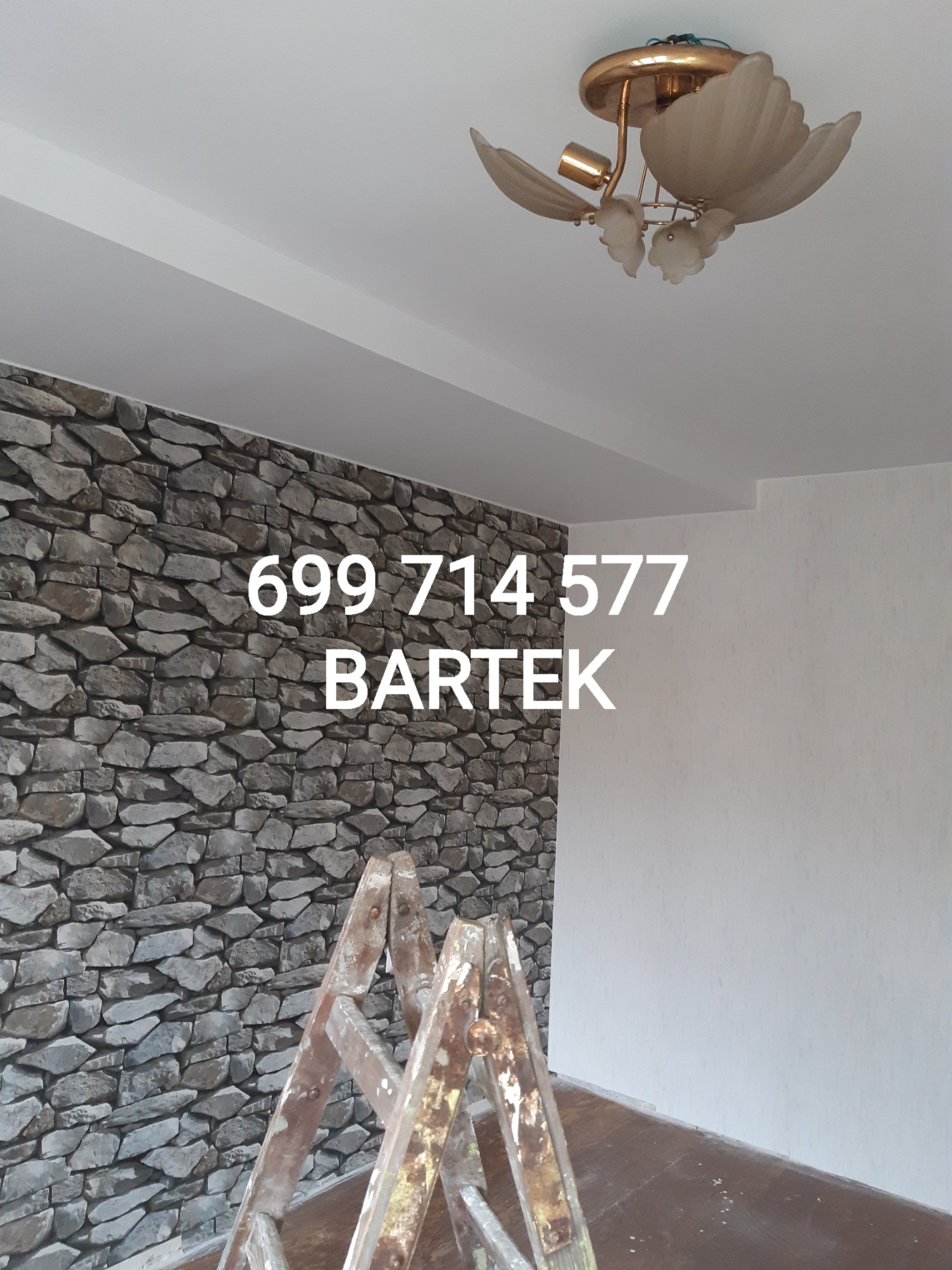 Ściana wykończona kamieniem dekoracyjnym w szarościach, widoczna drabina malarska, numer telefonu i imię wykonawcy naniesione cyfrowo na zdjęcie.