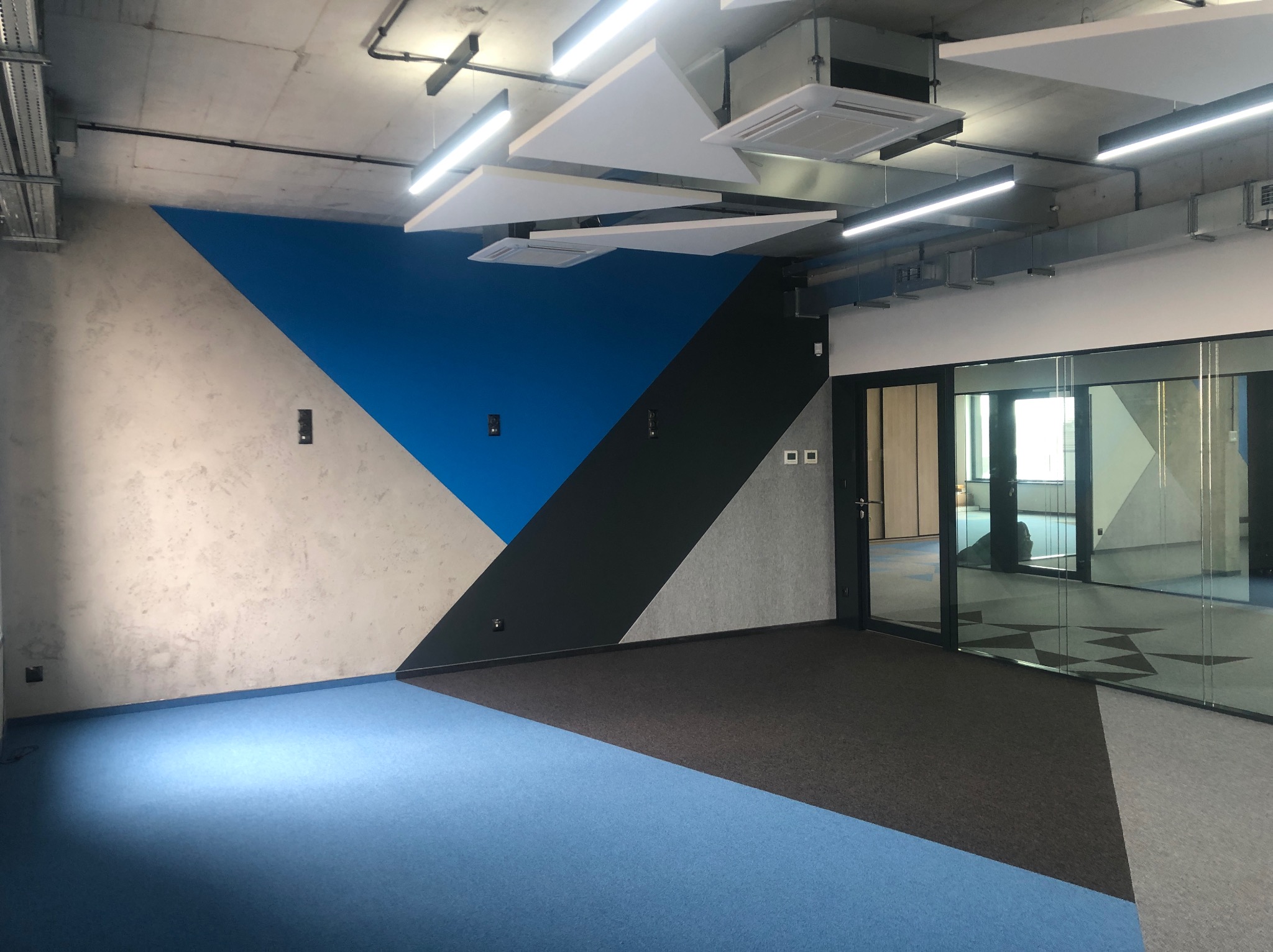 Wnętrze biura po remoncie: ściana z geometrycznym wzorem (błękit, szarość, czerń), sufit z elementami dekoracyjnymi. Podłoga z wykładziną w różnych odcieniach szarości i błękitu.