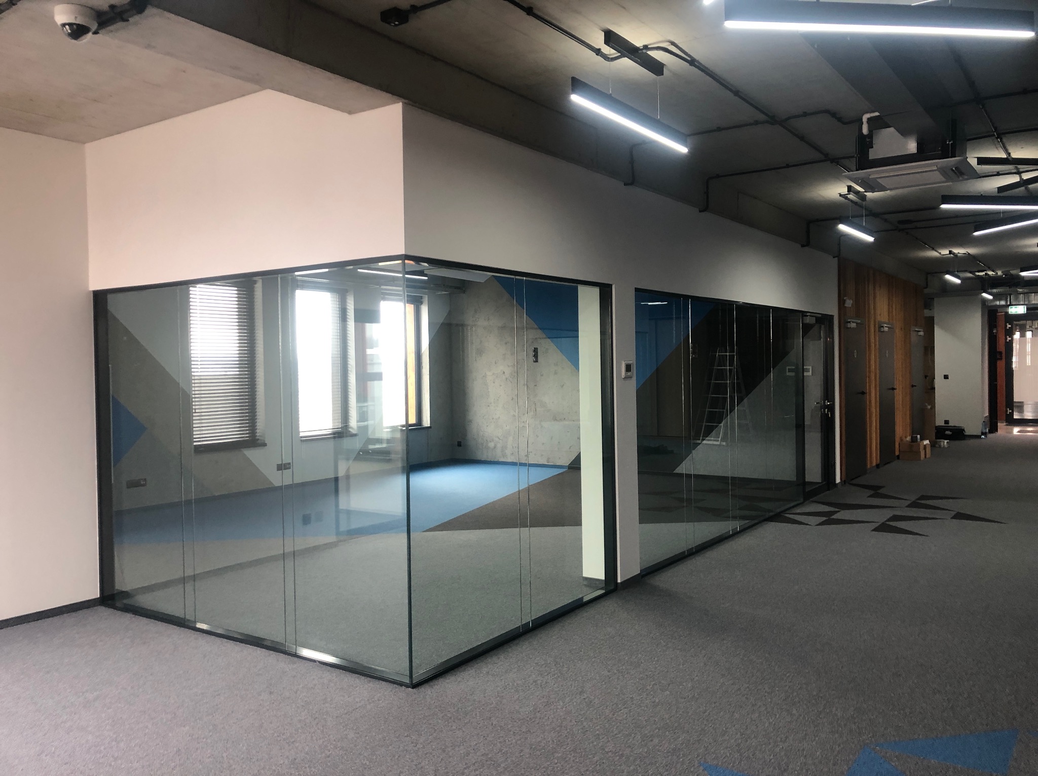 Szklana zabudowa biurowa z widoczną ścianą z betonu architektonicznego w tle. Widoczne elementy wykończenia wnętrz, oświetlenie sufitowe i szara wykładzina dywanowa.
