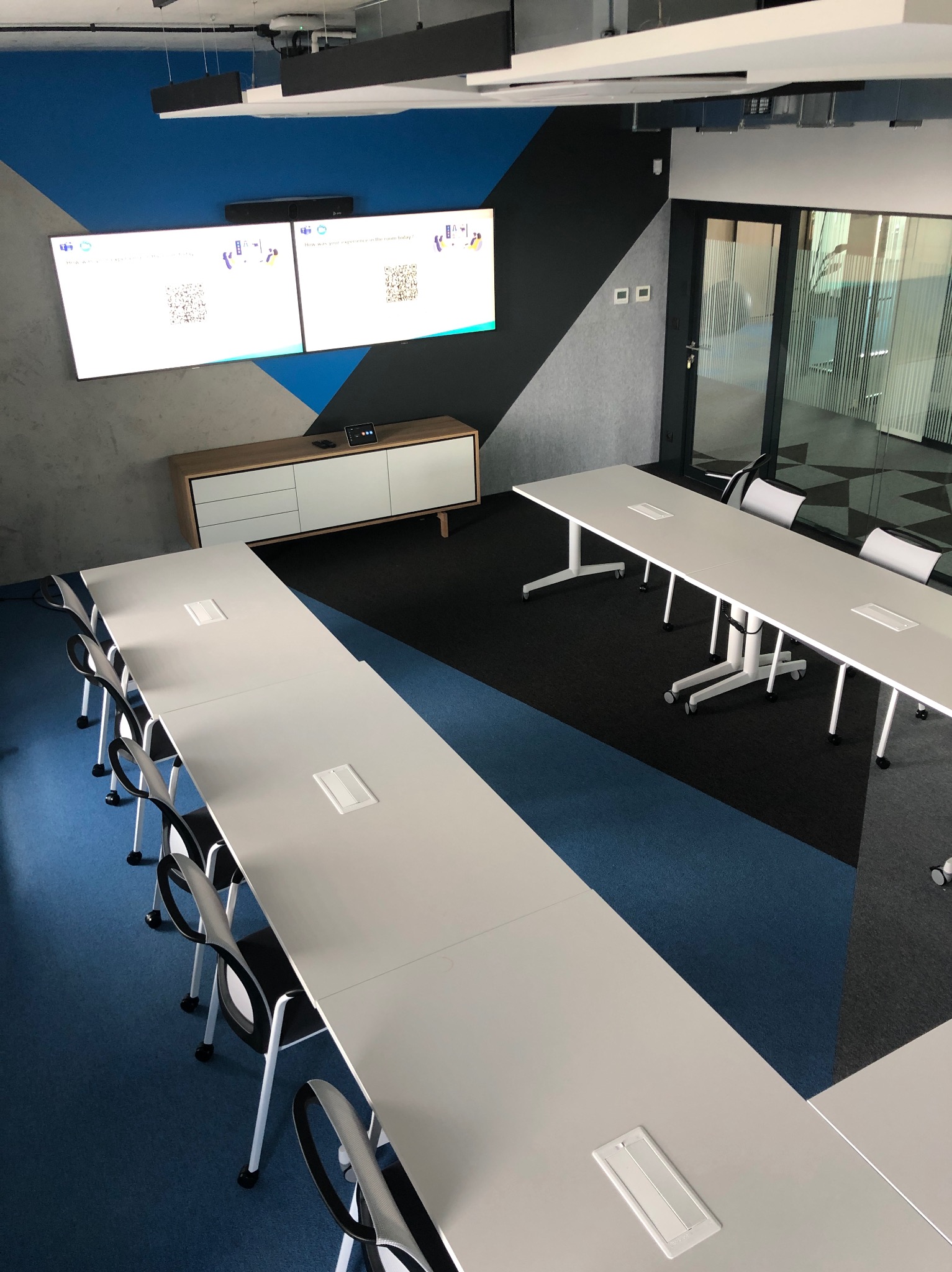 Nowoczesna sala konferencyjna po remoncie: długie białe stoły, czarne krzesła, ściana z geometrycznym wzorem w odcieniach szarości, czerni i błękitu, duży ekran.