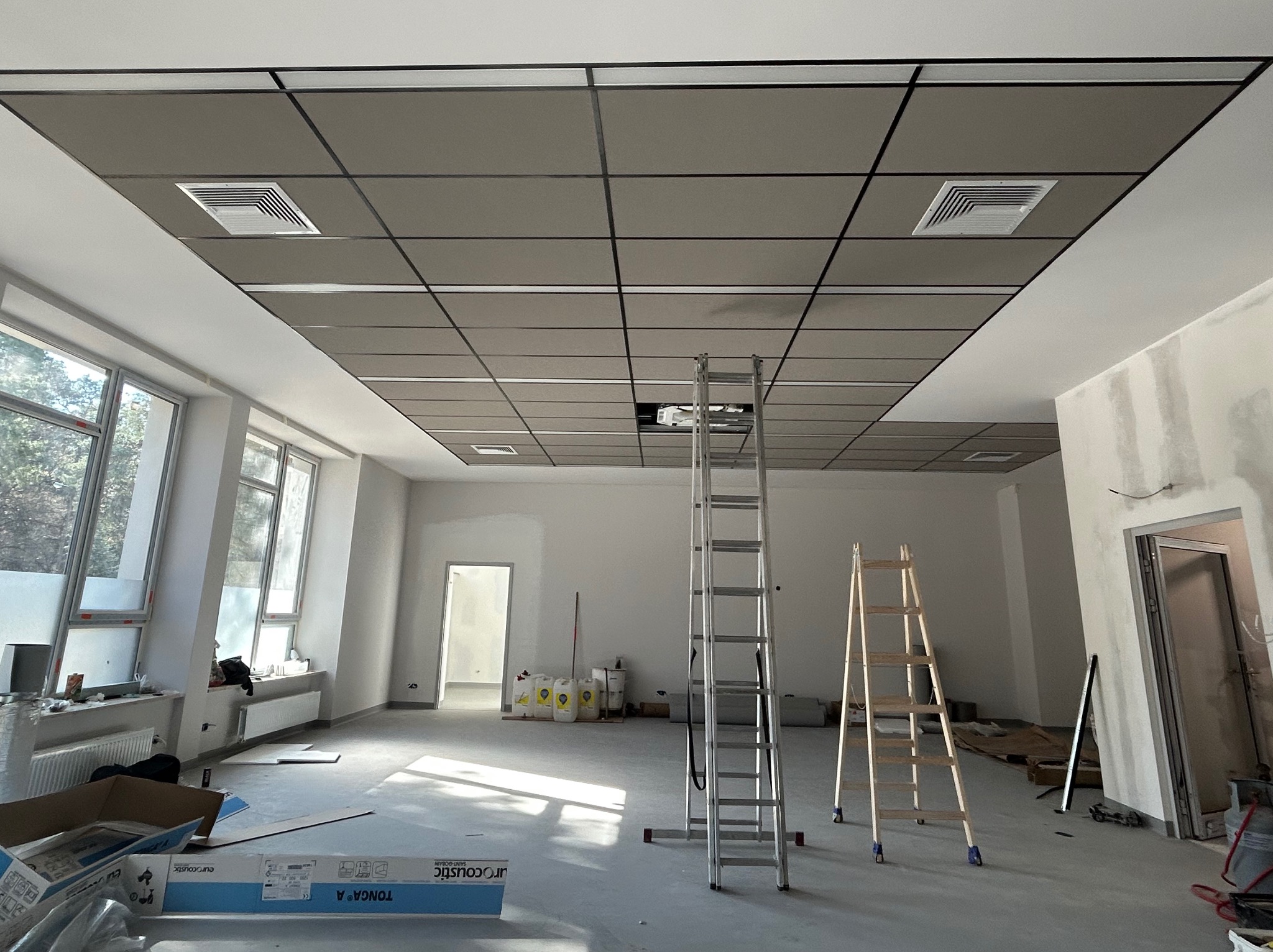 Montaż sufitu podwieszanego kasetonowego z kratownicą w przestronnym, jasnym pomieszczeniu biurowym. Widoczne drabiny i materiały budowlane.