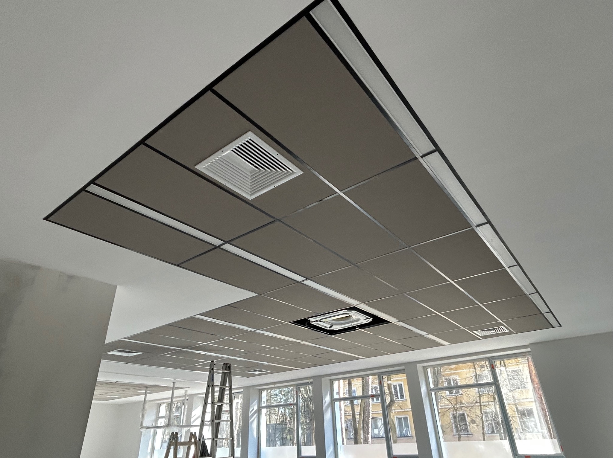 Sufit podwieszany kasetonowy w biurze, widok z dołu, kratki wentylacyjne, drabina oparta o ścianę, okna z widokiem na zewnątrz. Szare panele z czarną obwódką.