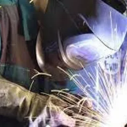 Pracownik w Dąbrowie Górniczej w kombinezonie ochronnym i masce spawalniczej wykonuje prace spawalnicze, z intensywnym snopem iskier oświetlającym stanowisko pracy.